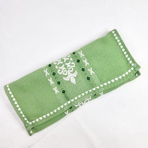 以下が含まれることがあります： 白の刺繍が施された、折り畳まれた緑色のリネンナプキン。ナプキンの中央には白い花の模様があり、小さな黒い点と白い「X」の形がアクセントになっています。縁は白いスカラップトリムで仕上げられています。