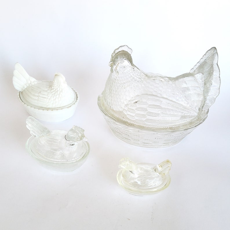 May include: Set of four clear glass chicken-shaped butter dishes. The largest dish features a chicken sitting in a nest. The other three dishes are smaller and feature a chicken without a nest.
