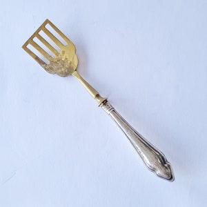 Puede incluir: Un pequeño utensilio de servicio con una cabeza de latón dorado de cuatro dientes rectangulares y un mango de metal plateado cónico. El mango presenta un diseño decorativo estriado y un collar distintivo donde se une al cuello de latón.