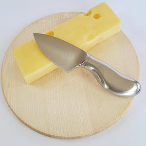May include: A block of yellow cheese with holes and a stainless steel cheese knife on a round wooden cutting board. The knife has a curved handle and a wide blade. The cheese and knife are set against a white background.