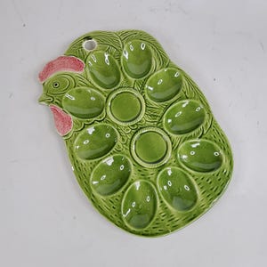 Könnte beinhalten: Grüne Eierservierplatte aus Keramik in Form eines Huhns. Die Platte hat 10 eiförmige und drei kreisförmige Vertiefungen. Der Hühnerkopf befindet sich auf der linken Seite, mit einem rosa Kamm und Kehllappen. Oben befindet sich ein Loch zum Aufhängen.