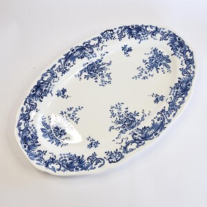 May include: An oval ceramic serving platter featuring a white base with a deep blue floral pattern. The rim is decorated with intricate scrollwork and foliage, while the center displays scattered clusters of flowers and leaves in blue.
