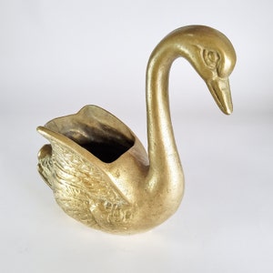 Pode incluir: Uma figura de cisne em latão polido com uma secção traseira oca. A superfície metálica apresenta um tom dourado quente com detalhes texturizados nas asas e um pescoço liso e curvo que se estende para cima e se inclina para a frente.