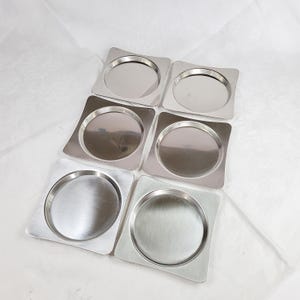 May include: A set of six square stainless steel coasters with rounded corners and a circular recessed centre. Each coaster features a polished metallic finish with a smooth, reflective surface, arranged in two columns of three on a white surface.