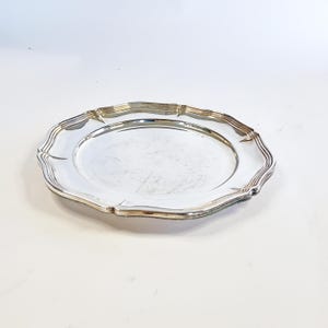 Può includere: Un piatto rotondo in metallo color argento con un centro liscio e riflettente e un bordo decorativo smerlato caratterizzato da molteplici creste concentriche. Il piatto ha una finitura lucida e un bordo ondulato che incornicia l'area centrale.