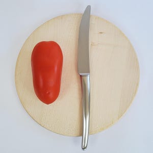 May include: A red Roma tomato and a stainless steel knife rest on a round wooden cutting board. The knife has a silver blade and handle. The tomato is smooth and oval-shaped. The cutting board is light brown.