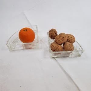 May include: Two clear glass, wedge-shaped dishes. One holds a bright orange tangerine. The other contains several brown walnuts. The dishes sit on a white surface.