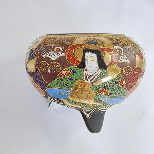 May include: A decorative, three-legged ceramic bowl with a detailed, hand-painted design. The bowl features a brown background with gold accents and a central figure in traditional Japanese attire. The figure is surrounded by intricate patterns in various colors.