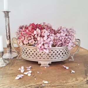 Peut inclure: Un panier en métal argenté avec un motif en treillis tressé et deux poignées ornées, rempli de fleurs d'hortensia séchées roses et mauves. Un grand bougeoir argenté élancé se trouve à gauche du panier sur une surface en bois.