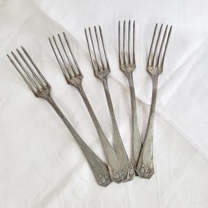 Può includere: Cinque forchette d'argento vintage con rebbi e manici decorativi sono disposte su una superficie bianca. Le forchette hanno un design classico, con un aspetto leggermente appannato, che suggerisce età e utilizzo. I manici presentano un motivo ornato.
