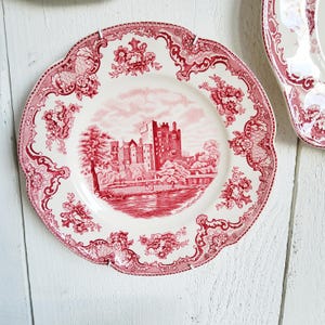 May include: A round ceramic plate with a scalloped edge, featuring a red and white transferware pattern. The center depicts a castle scene by a body of water, while the border is decorated with intricate floral motifs and scrollwork.