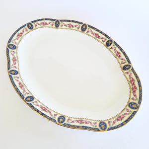 Könnte beinhalten: Eine ovale Servierplatte aus Keramik mit weißer Mitte und dekorativem Rand. Der Rand zeigt ein dunkelblaues Band mit goldenen Akzenten, unterbrochen von kleinen ovalen Medaillons mit gelben Blumenmotiven und Girlanden aus rosa Rosen.