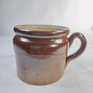 Könnte beinhalten: Ein zylindrischer Keramikbecher mit zweifarbiger brauner Glasur. Der obere Teil hat ein dunkleres, glänzendes Braun mit einem ausgeprägten Rand, während der untere Körper in einen helleren, matten Beigeton übergeht. Mit einem geschwungenen Henkel.