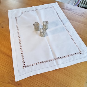 May include: White rectangular linen tablecloth with a decorative brown floral border. Three small, textured glass candle holders are centered on the tablecloth, which is placed on a wooden table.