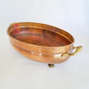 Può includere: Una bacinella ovale in rame con una finitura metallica lucida che presenta segni visibili di invecchiamento e patina. Il recipiente include due manici curvi in ottone fissati ai lati ed è sostenuto da quattro piccoli piedini in ottone.