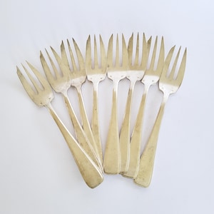 Op de afbeelding: Een set van zeven zilverkleurige metalen vorken in een waaiervorm. Elke vork heeft een kop met drie tanden en een glad, plat handvat dat naar de basis toe iets smaller wordt. Het metaal heeft een gepolijste afwerking met lichte gebruikssporen.