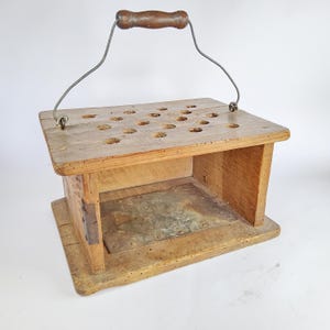 May include: A rectangular wooden box with a top surface featuring multiple circular holes, a metal wire handle with a wooden grip, and a metal base plate. The structure is constructed from light brown wood with visible grain and small dark surface markings.