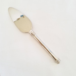 Puede incluir: Un servidor de pasteles de metal plateado con una hoja ancha, plana y puntiaguda, y un mango cilíndrico delgado. El mango presenta tres detalles de anillos horizontales cerca de la hoja y tres en la base. La hoja tiene una pequeña marca azul.