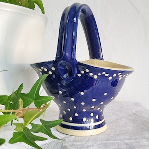 Può includere: Un cestino in ceramica blu scuro con un manico alto e arcuato e una base svasata. L'esterno presenta un motivo a piccoli pois bianchi sparsi sulla superficie, con una fascia bianca solida vicino alla base e un bordo interno color crema.