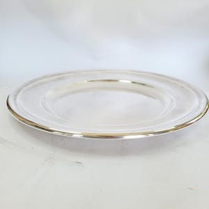 Op de afbeelding: Een ronde, zilverkleurige plaat met een iets verhoogde rand. De plaat heeft een gepolijst, reflecterend oppervlak en een dunne, goudkleurige band rond de rand. De plaat staat tegen een witte achtergrond.