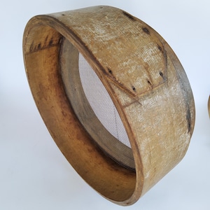 Puede incluir: Un tamiz de madera vintage con una malla fina. El tamiz tiene un marco circular de madera marrón claro, que muestra signos de desgaste y edad. La malla es de color gris claro. El tamiz se utiliza probablemente para tamizar harina u otros ingredientes secos.