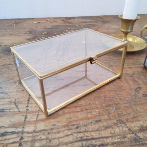 Puede incluir: Una caja expositora rectangular de cristal con un marco fino de latón y tapa abatible. Los paneles de cristal transparente están unidos por bordes de tono dorado, creando un recipiente de almacenamiento con un cierre sencillo al frente.