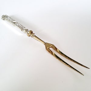 Pode incluir: Um garfo de entalhar prateado e dourado. O cabo é prateado com um design texturizado, enquanto os dentes do garfo são dourados. O garfo foi projetado para entalhar e servir carnes. O design geral é elegante e vintage.