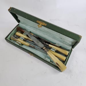 May include: A collection of antique knives with cream-coloured handles and silver-toned detailing, presented in an open green, textured case. The blades are silver and show signs of age. The case is lined with a pale blue fabric.