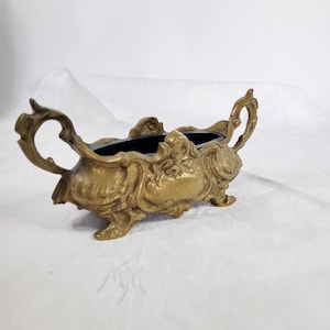 Puede incluir: Jardinera ornamentada en tono latón con interior negro. La jardinera presenta intrincados adornos y asas a cada lado. La pieza decorativa es probablemente vintage y adecuada para pequeñas plantas o arreglos florales.