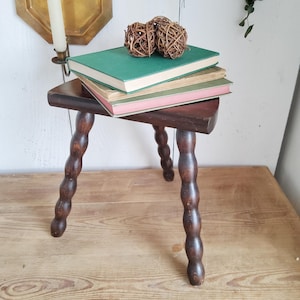 May include: A dark brown wooden stool with three twisted legs. The stool has a triangular top and is decorated with a stack of three books.
