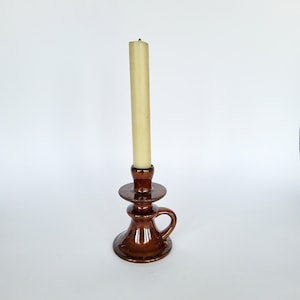 May include: A brown ceramic candlestick holder with a handle, holding a tall, off-white candle. The candlestick has a glossy finish and a classic design. The candle is unlit and stands upright in the holder. The overall aesthetic is vintage and decorative.