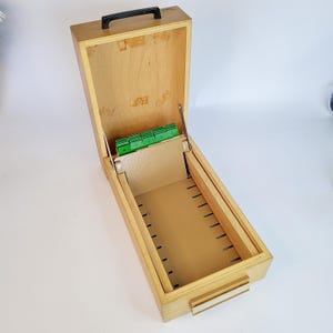 Könnte beinhalten: Offene Kartenkatalogbox aus Holz mit schwarzem Griff. Die Box ist hellbraun mit einem Klappdeckel und einer Reihe von Trennwänden mit den Buchstaben A bis D. Der Innenraum hat Schlitze für Karten. Auf der Vorderseite befindet sich ein kleiner, rechteckiger Etikettenhalter.
