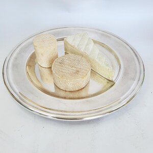 May include: A silver platter displays three cheeses: a cylindrical cheese, a round cheese, and a triangular wedge. The cheeses are pale, with textured surfaces. The platter has a wide, polished rim, creating a classic presentation.