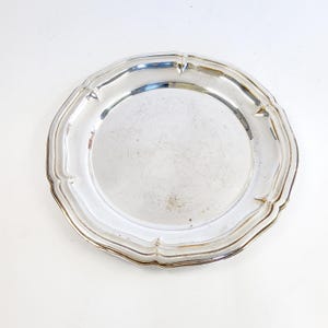 Può includere: Un piatto rotondo in metallo color argento con una superficie liscia e riflettente e un bordo ampio e smerlato con sottili rientranze. Il piatto presenta una finitura lucida con lievi graffi visibili sul centro e sui bordi.