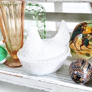 May include: White ceramic chicken-shaped butter dish with a woven basket design. The dish is sitting on a white wooden shelf with other decorative items.