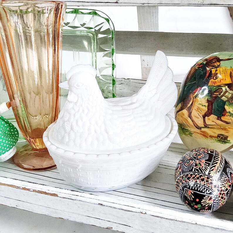 May include: White ceramic chicken-shaped butter dish with a woven basket design. The dish is sitting on a white wooden shelf with other decorative items.