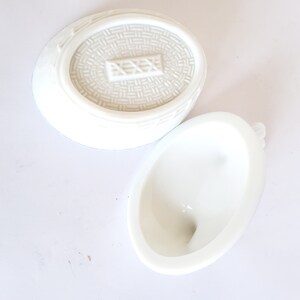May include: White ceramic egg-shaped container with a lid. The lid has a woven pattern and a raised rectangular design. The container is empty.