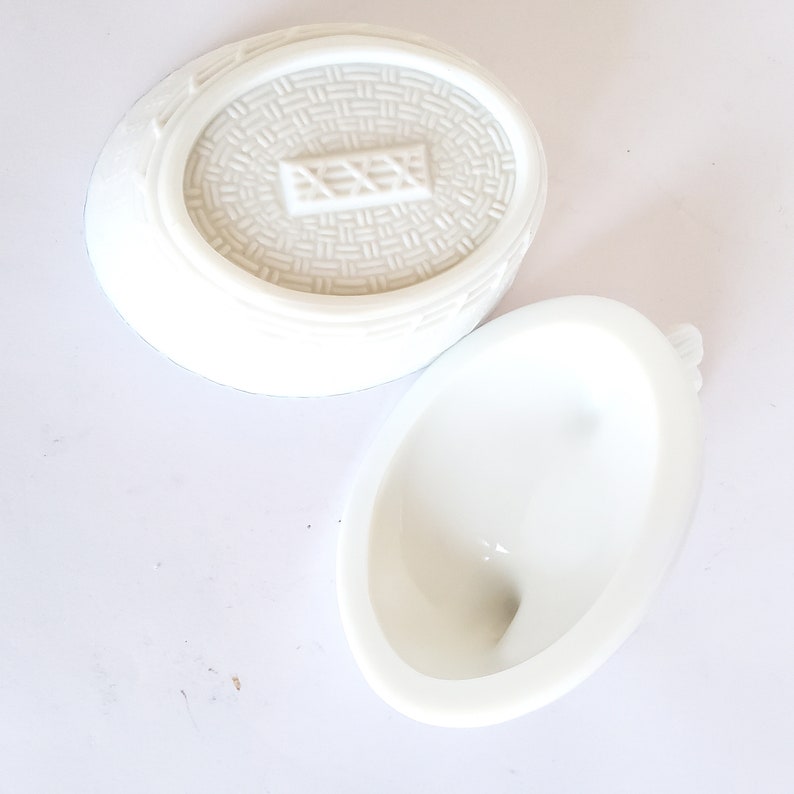 May include: White ceramic egg-shaped container with a lid. The lid has a woven pattern and a raised rectangular design. The container is empty.