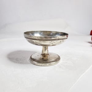 Puede incluir: Un pequeño cuenco antiguo con pedestal plateado. El cuenco tiene una base redonda, un tallo corto y un plato circular poco profundo. El borde del cuenco está decorado con una fina línea oscura. La base tiene un borde con cuentas.