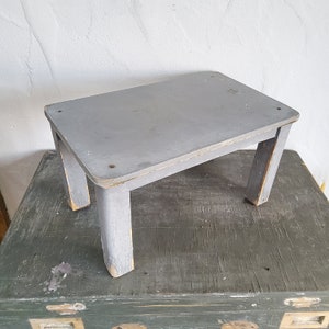 Könnte beinhalten: Ein kleiner, rechteckiger Holzhocker mit matter grauer Lackierung. Der Hocker hat vier stabile Beine und eine flache Oberseite mit sichtbaren Gebrauchsspuren, darunter abgeblätterte Farbe an den Kanten und Ecken, die das natürliche Holz darunter freilegen.