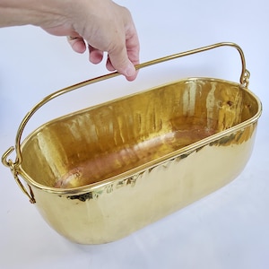 Puede incluir: Una jardinera de latón martillado con un asa curva. El recipiente de forma ovalada tiene un acabado dorado brillante. El asa está sujeta a los lados con pequeños soportes decorativos. La jardinera está sujeta por una mano.