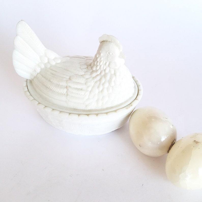May include: White ceramic chicken-shaped butter dish with a lid. The dish is decorated with raised details and has a scalloped edge. Two white ceramic eggs are beside the dish.