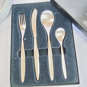 Puede incluir: Un juego de cubiertos de acero inoxidable, que incluye un tenedor, un cuchillo y dos cucharas, presentado en una caja negra. Los utensilios tienen un diseño sencillo y moderno con mangos redondeados y un acabado pulido.