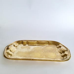 Könnte beinhalten: Ein rechteckiges, goldfarbenes Tablett mit verzierten Kanten. Das Tablett hat eine leicht gebogene Form und eine glatte, reflektierende Oberfläche. Die Kanten weisen dekorative Reliefmuster auf. Das Gesamtdesign ist vintage und elegant.