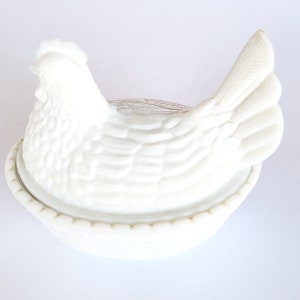 May include: White ceramic chicken shaped butter dish with a detailed design. The dish has a nest-like base and a lid that resembles a hen.
