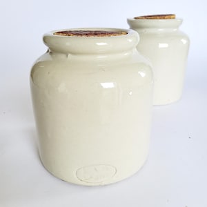 Op de afbeelding: Twee cilindrische voorraadpotten van gebroken wit keramiek met een gladde, glanzende afwerking. Elke pot heeft een dikke, afgeronde rand en is afgesloten met een platte, getextureerde bruine kurk. Onderaan is een ovaal merkteken zichtbaar.