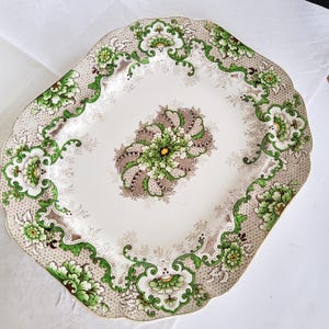 Könnte beinhalten: Eine ovale Servierplatte aus Keramik mit gewelltem Rand, die eine cremefarbene Basis mit komplizierten grünen und braunen Blumenmustern, geschwungenen Blattmotiven und einem zentralen, symmetrischen botanischen Designelement aufweist.