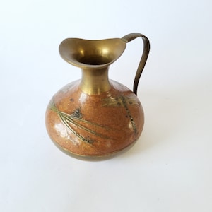 Pode incluir: Um pequeno jarro de latão com corpo arredondado e borda alargada. O corpo apresenta um acabamento texturizado em castanho manchado com motivos gravados de libélulas e ervas em verde e dourado. Inclui uma pega curva fixada ao pescoço.
