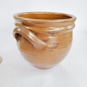 Può includere: Un vaso di ceramica marrone con corpo arrotondato e due manici. Il vaso ha un'ampia apertura e una superficie strutturata. Il colore è un marrone caldo e terroso, con striature più scure. Questo vaso fatto a mano è adatto per l'arredamento.