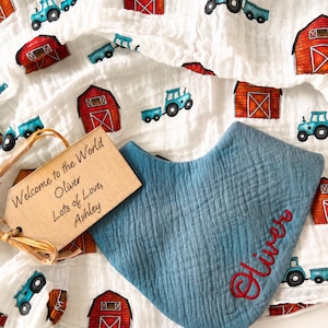 May include: A blue muslin bandana bib with the name Oliver embroidered in red thread, placed on a white muslin blanket featuring a repeating pattern of red barns and blue tractors, accompanied by a rectangular wooden tag reading Welcome to the World Oliver Lots of Love, Ashley.
